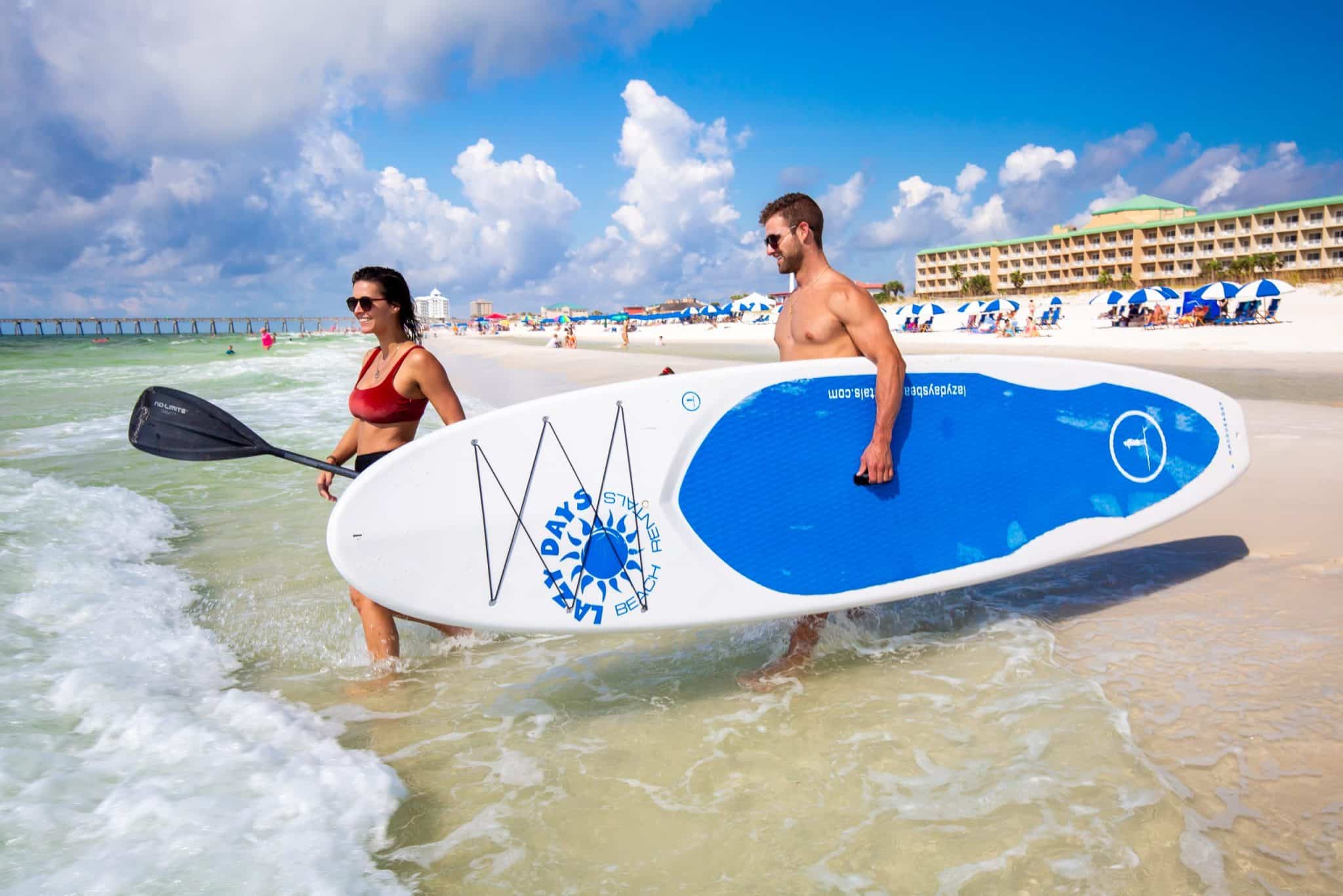 Man and woman walking into the tide at Pensacola Beach holding a paddle board.