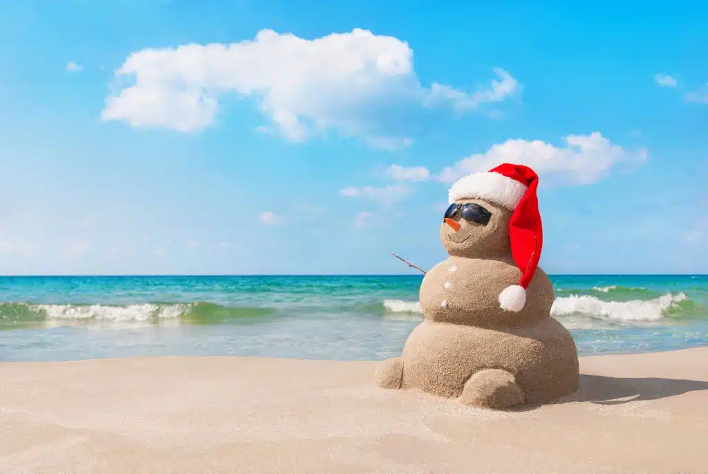 Sandy christmas snowman in red santa hat and sunglasses at sunny beach.